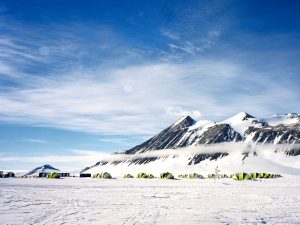 A row of yellow and green tents is set up on a snowy plain in Antarctica beneath a mountain with snow-covered peaks and a cloud band, under a bright blue sky with scattered clouds.
