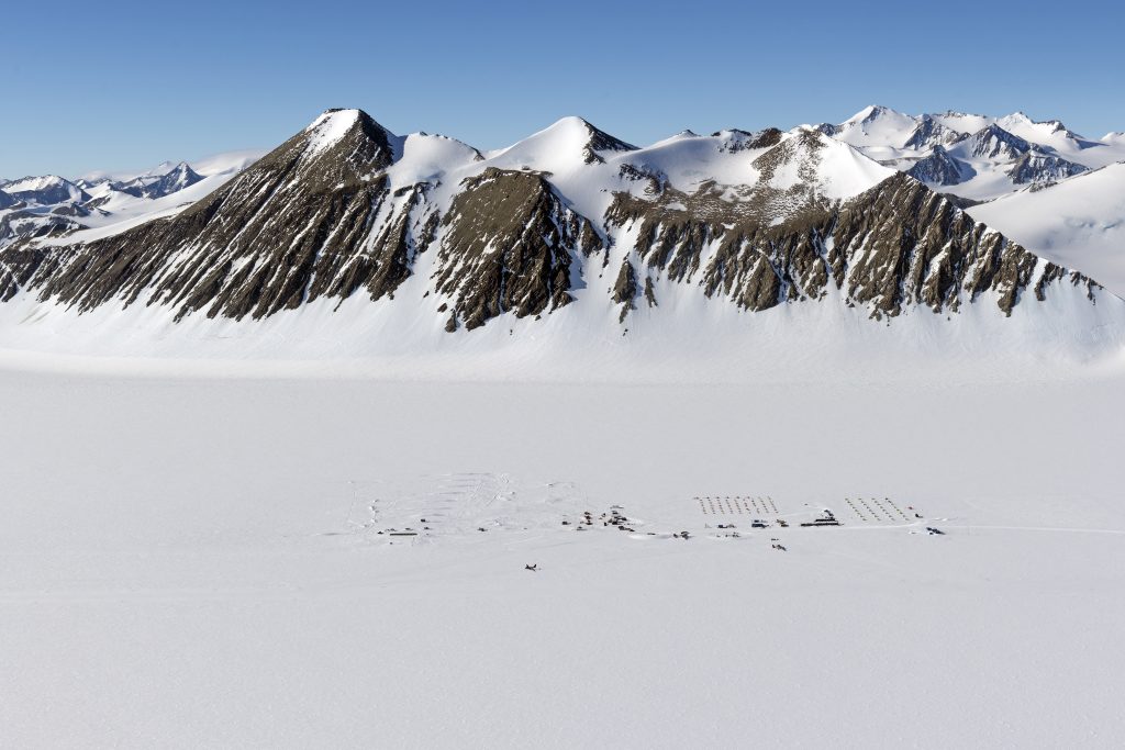 A remote scientific research camp is set up on a vast, snowy plain at the base of tall, rugged, snow-covered mountains under a clear blue sky in Antarctica.