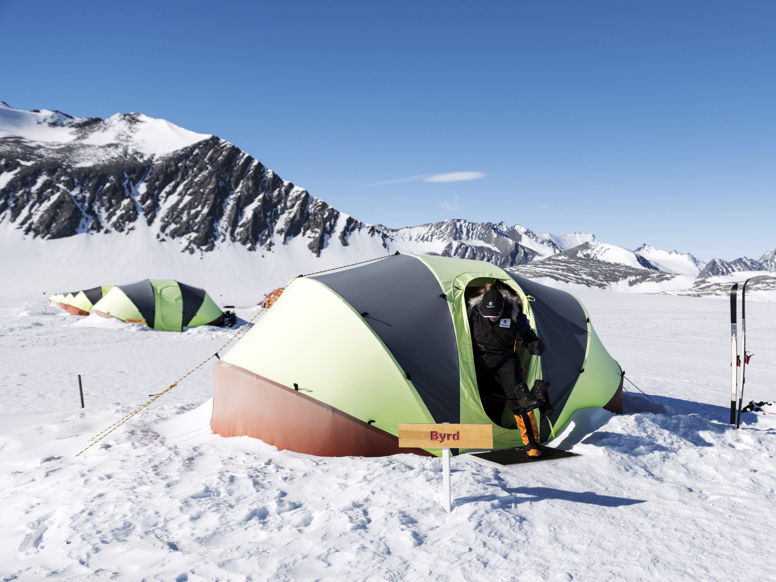 A person in cold-weather gear stands at the entrance of a green tent labeled Byrd, surrounded by snow at the South Pole in Antarctica, with mountains in the background and another Antarctic Logistics and Expeditions tent nearby under a clear blue sky.