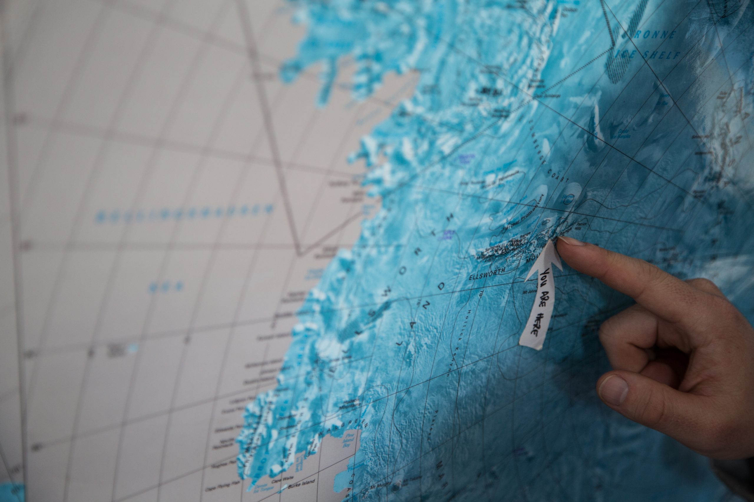 A hand points to a location labeled NY ÅLESUND on a blue-toned map, showing the coastline and geographic features of a northern region—reminiscent of maps used by Antarctic Logistics and Expeditions for polar exploration.
