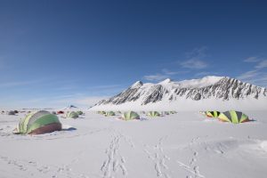 Multiple tents are set up on a snow-covered landscape with distant mountains under a clear blue sky. Footprints lead between the tents, revealing an Antarctic Logistics and Expeditions camp in remote Antarctica.