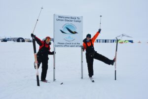 Two people in winter gear pose playfully with ski poles beside a sign reading Welcome Back to Union Glacier Camp! On Antarctica’s snowy terrain, with tents and buildings behind them, they celebrate a stop with Antarctic Logistics and Expeditions.