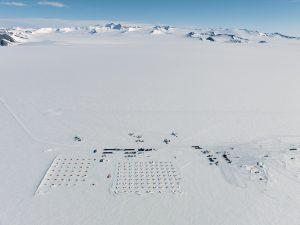 Aerial view of a snowy, remote Antarctic Logistics and Expeditions research camp in Antarctica, with rows of tents and several aircraft on a flat, white expanse, surrounded by distant mountains under a clear blue sky.