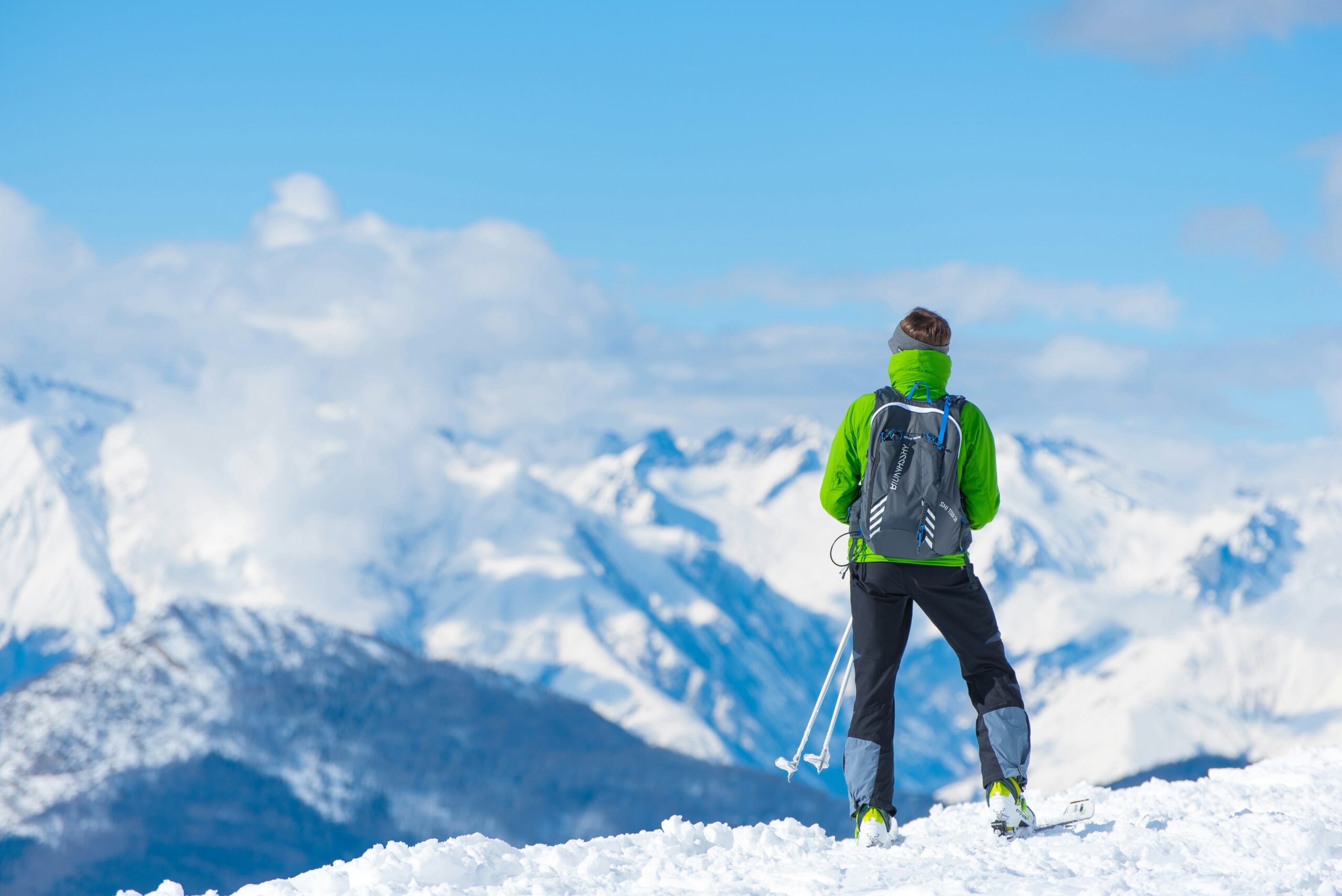 A person wearing a green jacket and black backpack stands on a snowy slope in Antarctica, facing a scenic view of snow-covered mountains near the South Pole under a clear blue sky, holding ski poles—an expedition made possible by Antarctic Logistics and Expeditions.