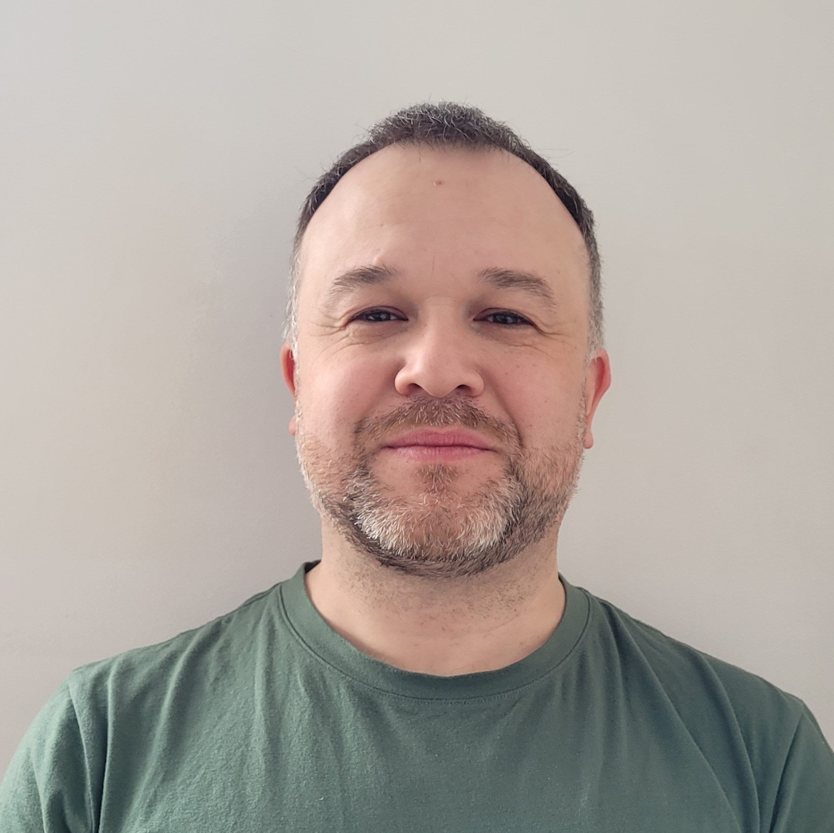 A man with short dark hair and a beard is smiling softly at the camera. Wearing a green T-shirt, he stands in front of a plain light-colored wall, reminiscent of photos taken by explorers at Antarctic Logistics and Expeditions near the South Pole.