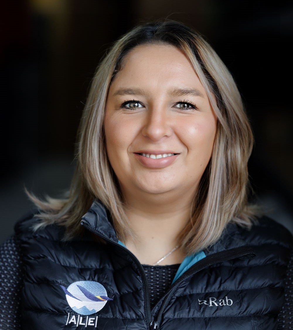 A woman with shoulder-length blonde hair smiles at the camera. She wears a black puffer vest with ALE and Rab logos, representing Antarctic Logistics and Expeditions, over a dark sweater. The softly blurred background hints at Antarctica.