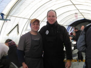 Two men smiling and posing inside a large, white, tent-like structure in Antarctica with several people sitting at tables in the background. One man wears an apron; both are dressed warmly. The timestamp reads 11/01/2009 14:38.