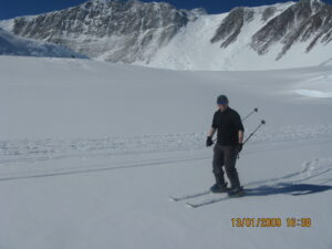A person wearing dark winter clothing and sunglasses skis on a snowy mountain slope near rugged peaks in Antarctica. The sky is clear and blue, and ski tracks are visible in the snow—a true adventure with Antarctic Logistics and Expeditions.