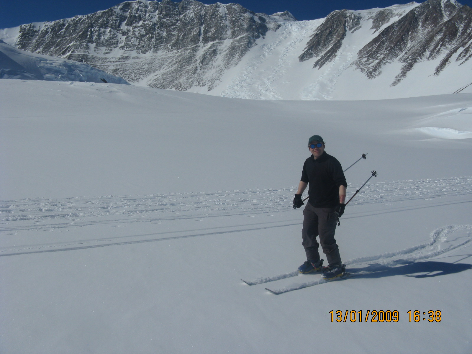 A person wearing dark winter clothing and sunglasses skis on a snowy mountain slope near rugged peaks in Antarctica. The sky is clear and blue, and ski tracks are visible in the snow—a true adventure with Antarctic Logistics and Expeditions.