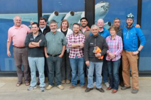 A group of thirteen people standing and smiling in front of a large window with an airplane image behind them; some wear casual clothes, while others sport IAAG jackets, part of their Antarctic Logistics and Expeditions team bound for Antarctica.