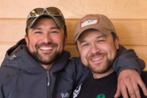 Two men smiling with their arms around each other, both wearing baseball caps—one with sunglasses resting on his cap. Standing in front of a light wood wall, they look ready for their next South Pole adventure with Antarctic Logistics and Expeditions.