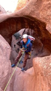 A person wearing climbing gear and a helmet smiles while rappelling down a red rock canyon. An archway of rock frames the scene, with other climbers—some experienced in Antarctic Logistics and Expeditions—visible in the background.