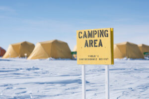 Yellow tents stand on a snowy landscape under a clear blue sky in Antarctica. A yellow sign in the foreground reads Camping Area – World's Southernmost Resort.