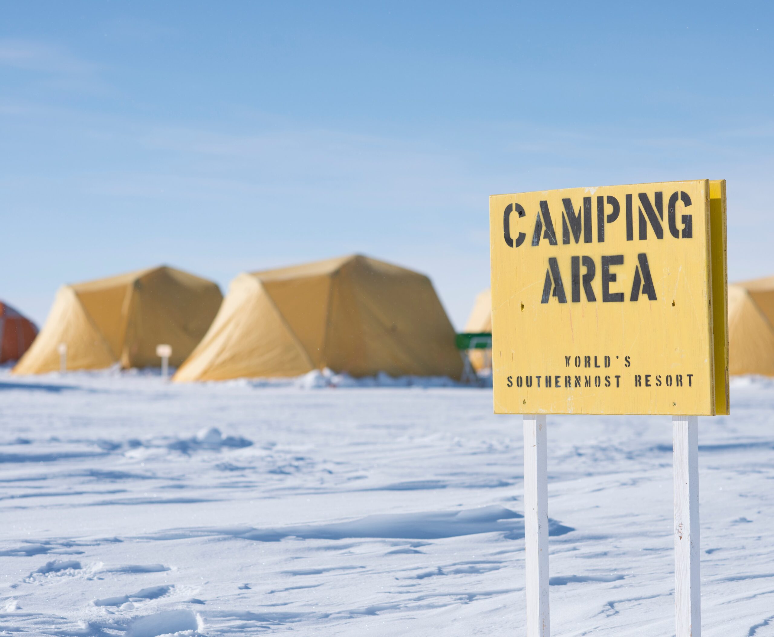 A yellow sign reading CAMPING AREA: WORLDS SOUTHERNMOST RESORT stands in a snowy Antarctic landscape with several yellow tents set up in the background under a bright blue sky.