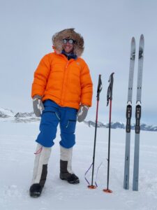 A person in a bright orange jacket, blue pants, and snow boots stands on a snowy landscape in Antarctica next to two ski poles and skis. Snowy mountains and a cloudy sky are visible in the background, evoking an Antarctic adventure.