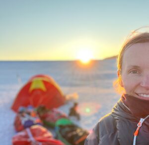 A person in cold weather gear smiles at the camera with a snowy Antarctic landscape, a sled with supplies, and the sun setting in the background during an expedition supported by Antarctic Logistics and Expeditions.