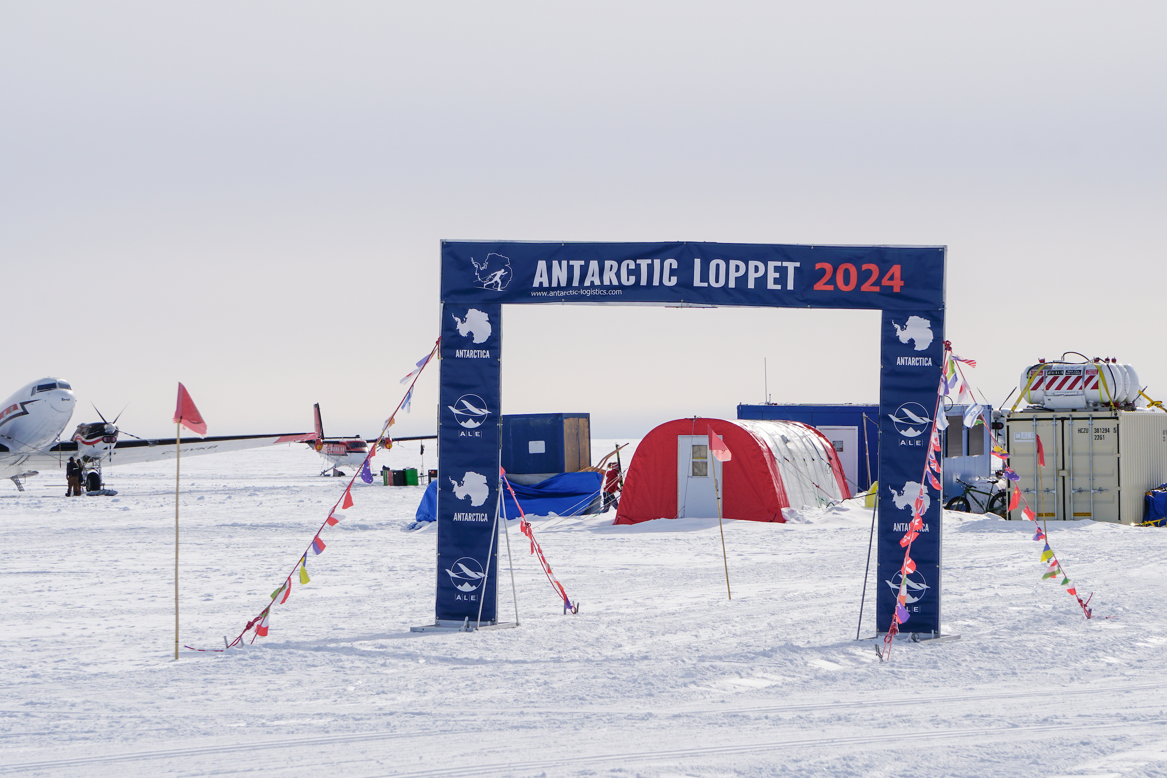 A snowy scene in Antarctica shows a race archway labeled “Antarctic Loppet 2024.” Behind it are colorful tents, flags, and an airplane set up by Antarctic Logistics and Expeditions on the ice under a pale sky.