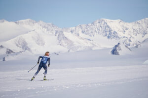 A cross-country skier in a blue outfit with the number 5 skis across a snowy landscape near towering, snow-covered mountains under a clear blue sky—experiencing the thrill of Antarctica with Antarctic Logistics and Expeditions.