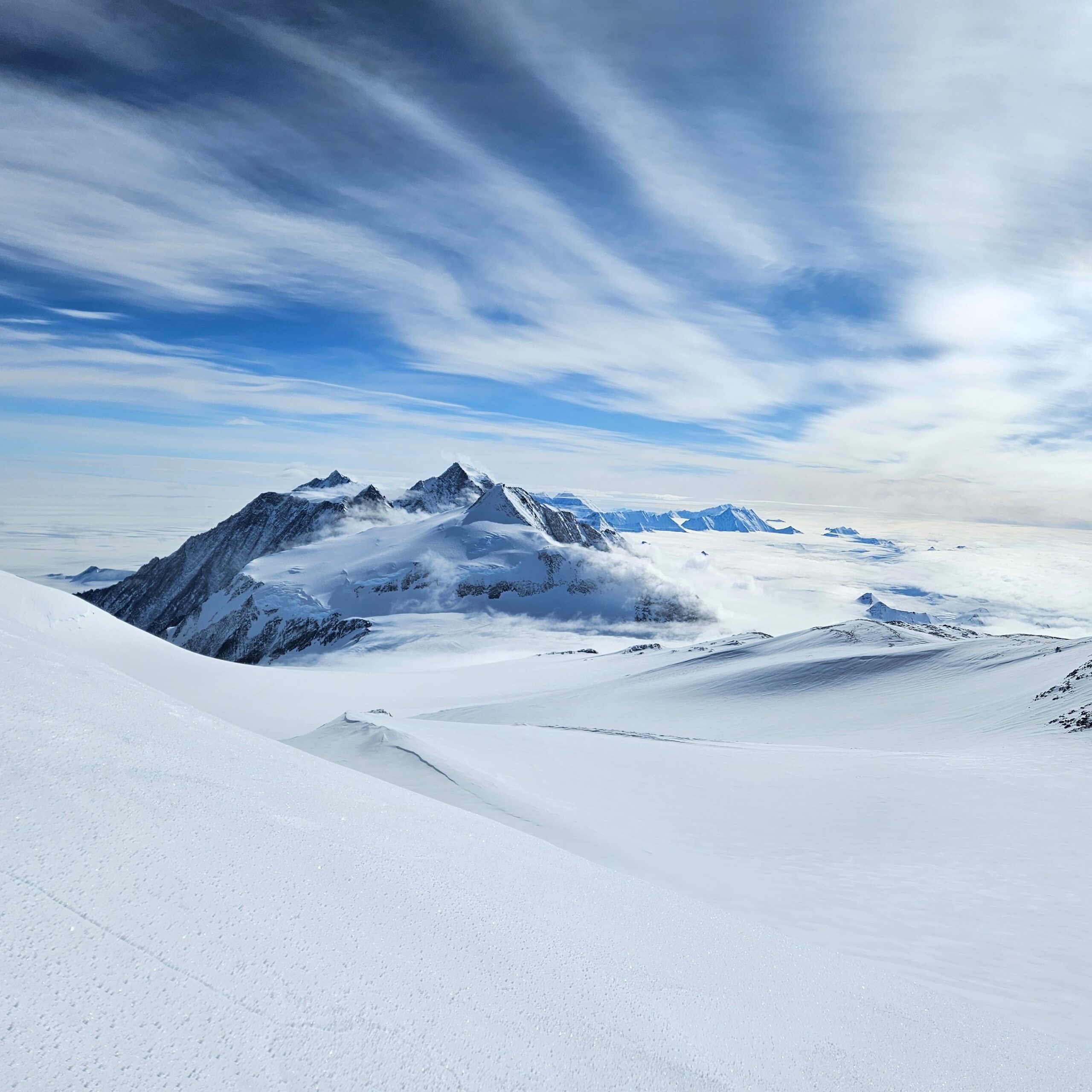 Snow-covered mountain peaks rise above a vast, icy landscape in Antarctica under a dramatic, partly cloudy sky with streaks of blue and white—a cold, remote, and pristine scene ideal for Antarctic Logistics and Expeditions.