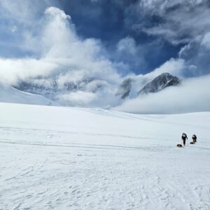 Two people trek across a vast snowy landscape in Antarctica toward towering, cloud-covered mountains under a dramatic sky, each pulling a sled over the pristine snow.