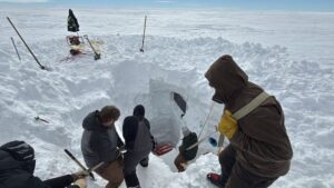 Several people wearing winter clothing work together to excavate a deep snow pit in the vast, snowy landscape of Antarctica. Tools and equipment from Antarctic Logistics and Expeditions lie nearby, with a sled at the edge of the pit.
