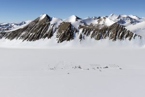 A remote scientific research camp, set up by Antarctic Logistics and Expeditions, sits on a vast, snowy plain at the base of rugged, snow-covered mountains in Antarctica beneath a clear blue sky. The camp structures appear small against the dramatic landscape.