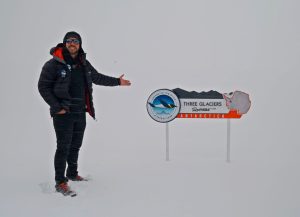 A person in winter clothing stands on snow, smiling and gesturing toward a sign that reads Three Glaciers, Antarctica in a snowy, foggy landscape.