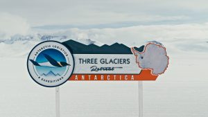 A sign in snowy Antarctica reads “Three Glaciers Retreat, Antarctica,” with a map outline of the continent and a logo for Antarctic Logistics & Expeditions against a backdrop of snow and distant mountains.