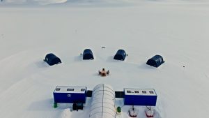 A snowy landscape with five black tents arranged in a semicircle around a wooden table and chairs, near two blue rectangular buildings and a white tunnel-like structure.