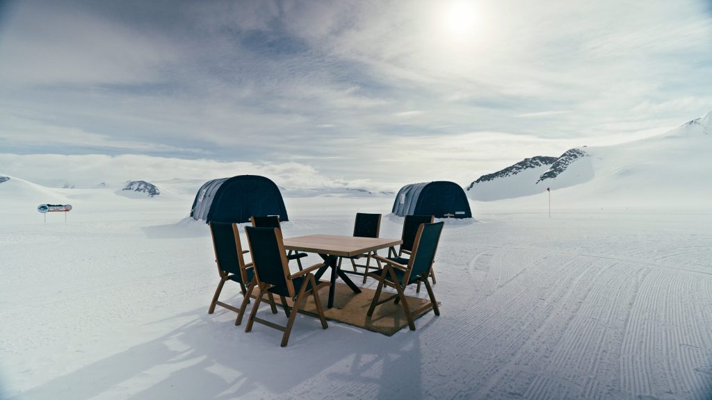A table with four chairs sits on a snowy landscape in front of two dark tents, with mountains and a cloudy sky in the background. The scene is bright, with sunlight reflecting off the snow.