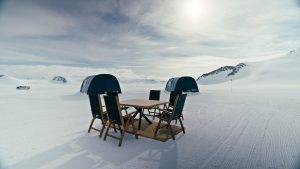 A table with four chairs sits on a snowy landscape in front of two dark tents, with mountains and a cloudy sky in the background. The scene is bright, with sunlight reflecting off the snow.