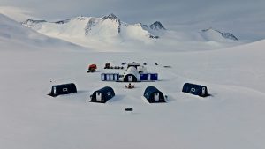 A remote research camp with several blue and white tents and modular buildings set up on snowy ground, surrounded by mountains under a cloudy sky in a polar landscape.