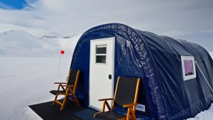 A blue arched tent with a white door and two wooden chairs sits on a snowy landscape, with mountains in the background and a red flag next to the tent.