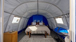 Interior of a cozy glamping tent with a double bed, beige bedding, a striped bench, wooden nightstand, rug, and blue jacket on a chair. Windows on both sides let in natural light.