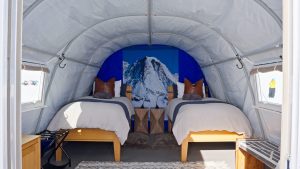 A cozy tent-like room with two single beds, wooden frames, brown pillows, and a large wall mural of a snowy mountain behind the beds. The room is bright and has modern decor with simple furnishings.