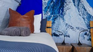 A cozy bed with a brown leather pillow, white and gray pillows, and rolled gray towels sits beside two wooden nightstands. The wall features a large, vibrant image of snowy mountain peaks.