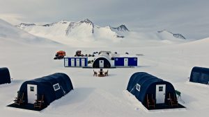 A snowy camp with blue tents, portable toilets, and a few trucks sits in a vast, white landscape surrounded by snow-covered mountains under a cloudy sky.