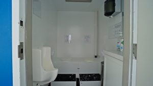 A small, white restroom with a urinal on the left, two toilet seats in the back, a sink and soap dispenser on the right, and signs on the wall above the toilets. The room appears clean and functional.