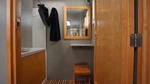 A small, modern bathroom with wood accents, a sink with a counter, a mirror above, a wooden stool, and a shower area with a black curtain. The floor is wooden slats and the door is partially open.