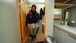 A man in winter clothing stands in a small wooden shower stall holding a large metal bucket, inside a modern bathroom with white counters and shelves.