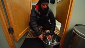 A person in warm clothing crouches indoors, filling a large metal pot with water from a small tap near the floor, surrounded by wooden panels and a trash can.