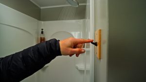 A person wearing a dark jacket presses a black switch on a beige wall near a shower, with water running from the showerhead in the background.