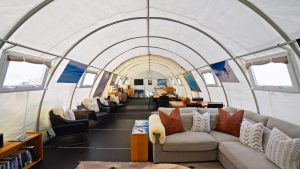 A cozy, modern lounge inside a white tent structure with armchairs, a grey sectional sofa, bookshelves, and large windows, decorated with photos and furnished for comfort.