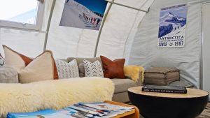 A cozy tent interior with a sofa covered in neutral pillows and a fur throw, a round coffee table with books, ottomans, and posters of Antarctica on the walls, creating a comfortable, adventure-themed space.