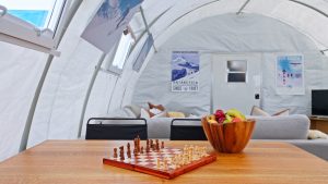 A chessboard and a wooden bowl of fruit sit on a table inside a bright, modern tent with couches and Antarctica-themed posters on the walls in the background.