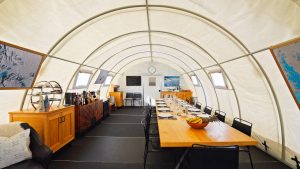 Interior of a bright, curved, tent-like dining space with a long wooden table set for a meal. There’s a buffet and bar along one wall, framed photos, windows, and a cozy sitting area on the left.