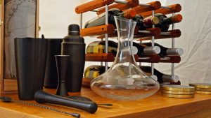 A wooden wine rack holds several bottles next to a clear glass decanter, cocktail shaker, black bar tools, coasters, and a map, all arranged on a wooden surface.