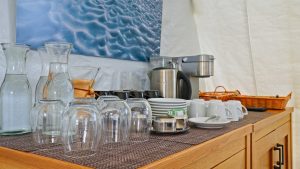 A countertop holds glass pitchers, upside-down drinking glasses, cups, saucers, a kettle, a coffee maker, and a basket, with a blue abstract painting hanging on the wall behind them.