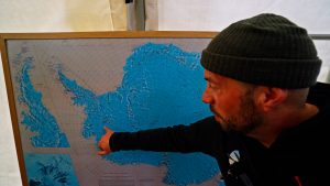 A man wearing a dark beanie points at a map of Antarctica, focusing on a region along the coastline, inside a tent or enclosed space.