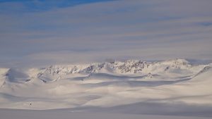 Snow-covered mountains under a partly cloudy blue sky, with smooth white slopes and jagged peaks in the distance, creating a serene winter landscape.